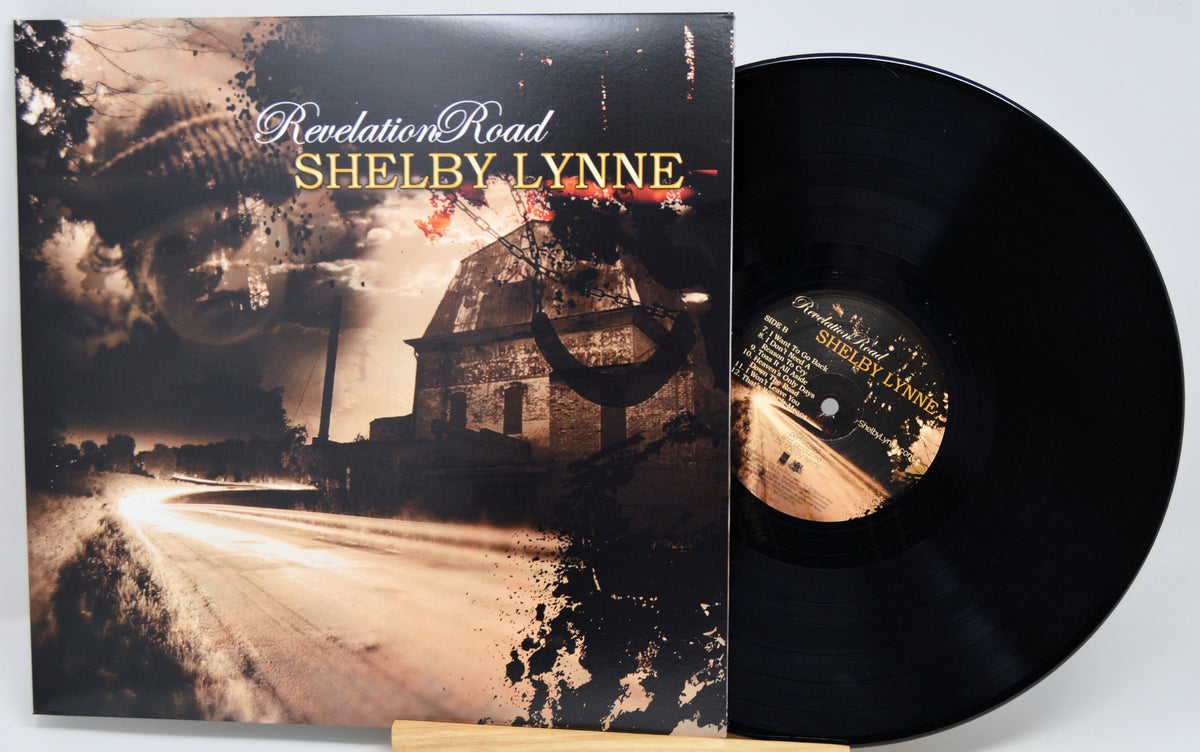 Vinyl record album 'Revelation Road' by Shelby Lynne, showing the illustrated black-and-brown album cover standing beside the black LP disc on a small wooden stand against a light gray background.