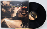 Vinyl record album 'Revelation Road' by Shelby Lynne, showing the illustrated black-and-brown album cover standing beside the black LP disc on a small wooden stand against a light gray background.