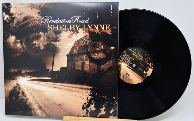 Vinyl record album 'Revelation Road' by Shelby Lynne, showing the illustrated black-and-brown album cover standing beside the black LP disc on a small wooden stand against a light gray background.