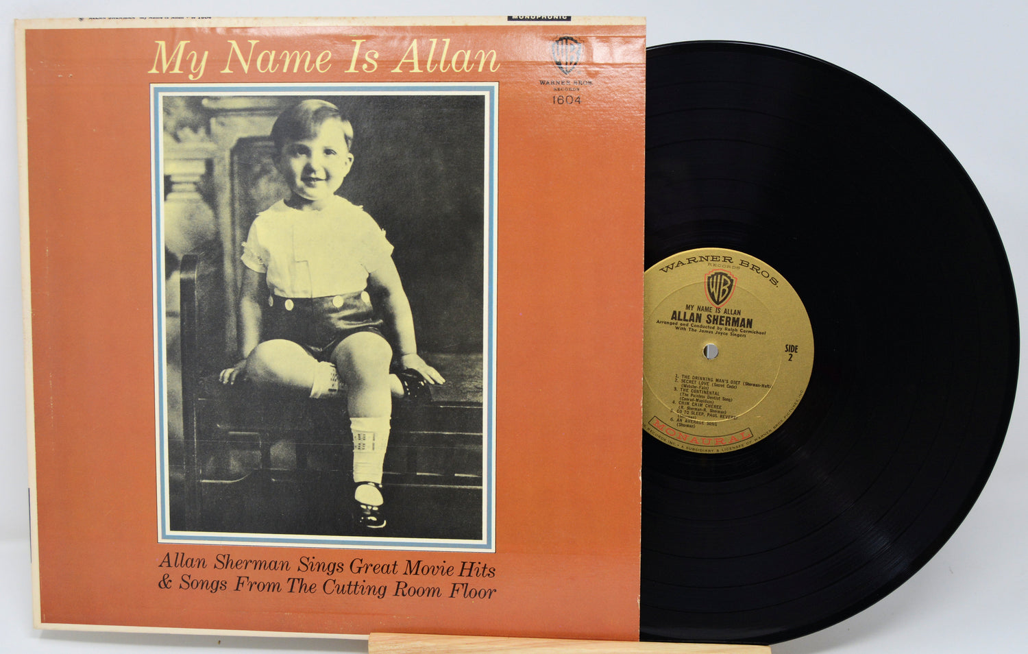 Vinyl record with orange cover featuring a black and white photo of a child, on a light gray background.