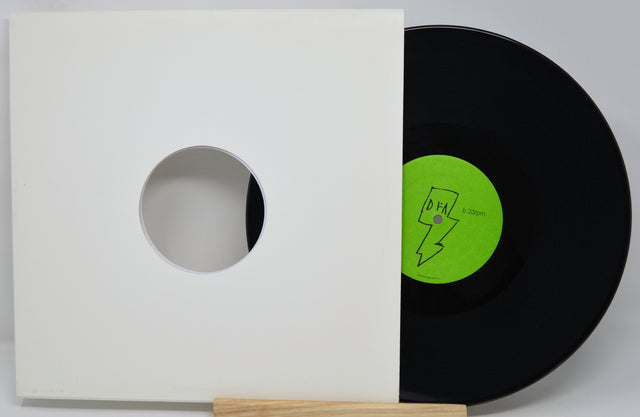 A black vinyl record partially pulled from a white square inner sleeve with a circular cutout, resting on a small wooden stand against a light gray background. The record label is bright green with a lightning-bolt graphic and the text 'DFA'.