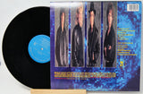 A vinyl record partially pulled from its sleeve, showing the black record with a blue center label and the back cover featuring four portrait photos of a band against a blue pixelated background.