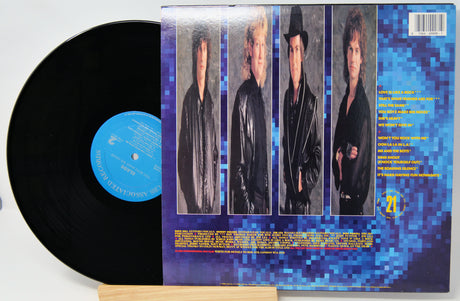 A vinyl record partially pulled from its sleeve, showing the black record with a blue center label and the back cover featuring four portrait photos of a band against a blue pixelated background.