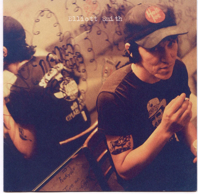 Elliott Smith album cover for 'Either Or' featuring a black and white photograph of Elliott Smith wearing a cap, sitting in front of a graffiti-covered wall.