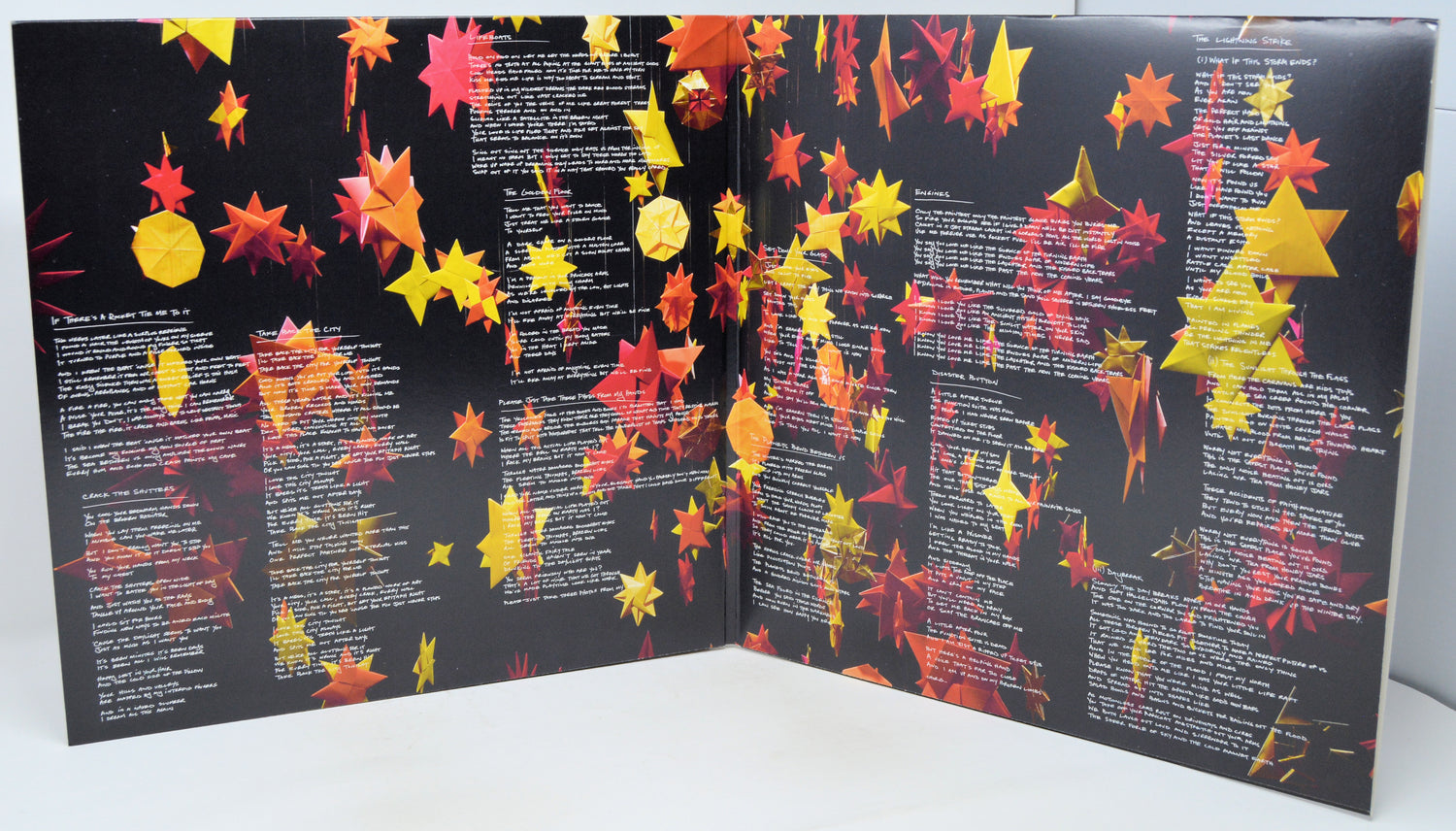 An open book or booklet with black pages covered in rows of white text and decorated with many hanging red, orange, and yellow paper-like leaves, photographed against a plain light background.
