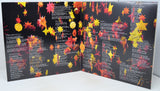 An open book or booklet with black pages covered in rows of white text and decorated with many hanging red, orange, and yellow paper-like leaves, photographed against a plain light background.