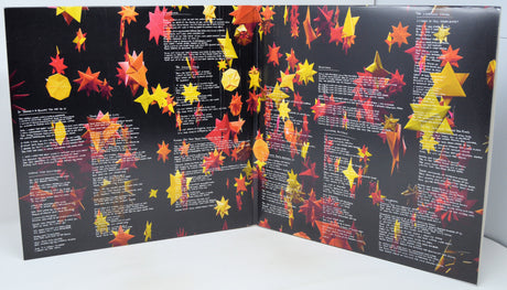 An open book or booklet with black pages covered in rows of white text and decorated with many hanging red, orange, and yellow paper-like leaves, photographed against a plain light background.