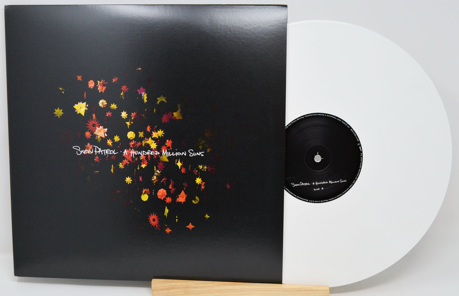 A vinyl record partially pulled from its sleeve, shown on a light wood stand against a white background. The sleeve is black with a colorful scattered floral or starburst graphic in red, orange, yellow, and white text.
