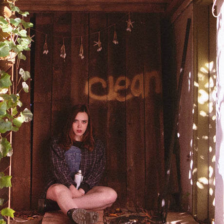 A person sitting cross-legged against a wooden backdrop with the word 'clean' painted on it, surrounded by foliage and hanging decorations.
