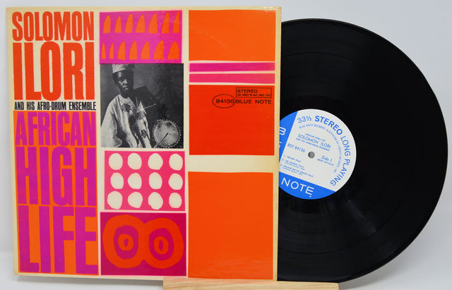 Vinyl record with a colorful cover featuring text and images, on a light gray background.
