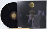 A vinyl record partially pulled from its black outer sleeve, showing the back of the package with tracklist text and a gold circular logo. The record and sleeve are photographed against a plain light background.