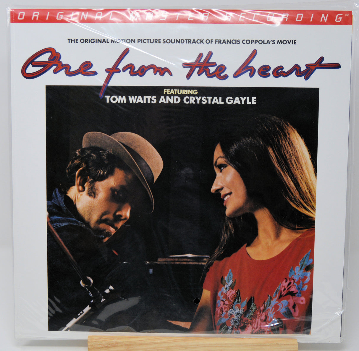 Vinyl record album cover for 'One from the heart' soundtrack, featuring a photo of Tom Waits and Crystal Gayle, sealed in plastic shrink wrap and displayed on a small wooden stand.