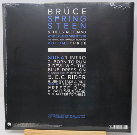 The back cover of a Bruce Springsteen & The E Street Band vinyl record, titled 'Winterland Night 1978: The Classic San Francisco Broadcast Volume Three'. The cover is black with white and blue text, wrapped in clear plastic, and photographed resting on a light-colored wooden stand.