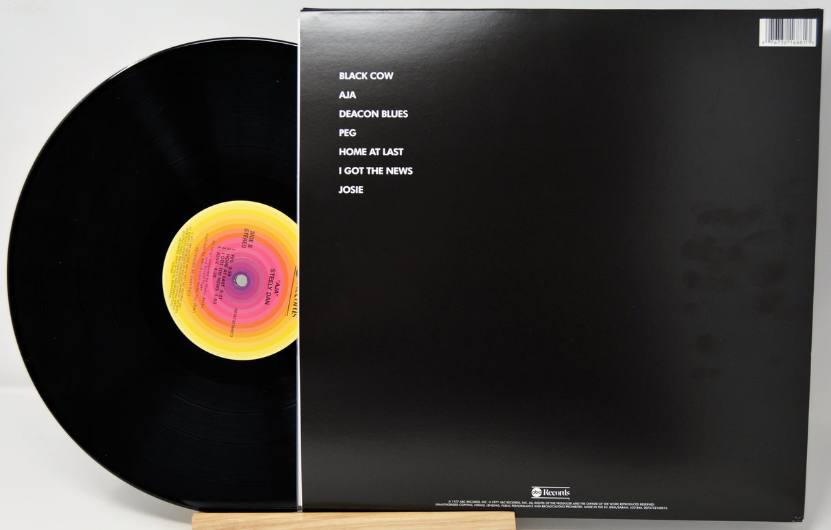 A vinyl record partially pulled from its black sleeve, photographed on a light background. The record is black with a yellow, pink, and orange center label.