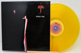 Steely Dan's 'Aja' vinyl record album with the black cover jacket and a translucent yellow record partially visible behind it on a white background.