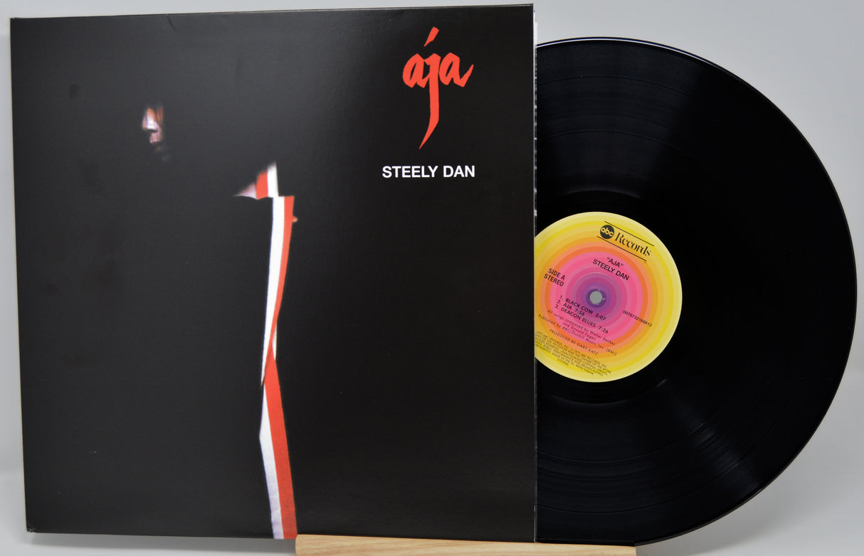 Steely Dan 'Aja' vinyl record LP with the black album cover partially slid off, revealing the black record and a yellow-and-pink center label, photographed on a light gray background.