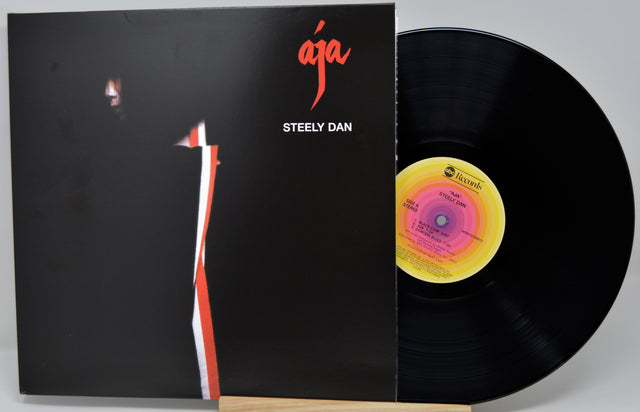 Steely Dan 'Aja' vinyl record LP with the black album cover partially slid off, revealing the black record and a yellow-and-pink center label, photographed on a light gray background.