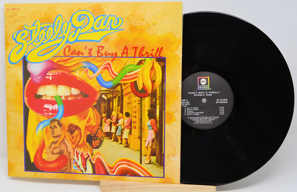 A vinyl record and its illustrated album cover for Steely Dan's 'Can't Buy A Thrill', displayed on a small wooden stand against a light gray background.