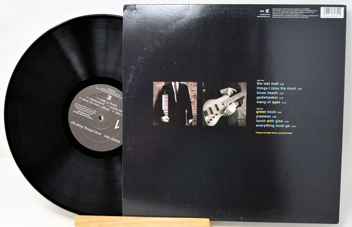 A vinyl record partially pulled from its black outer jacket, displayed on a small wooden stand against a white background. The back cover shows two small square photos and a track list printed in white and yellow text.