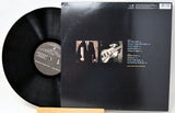 A vinyl record partially pulled from its black outer jacket, displayed on a small wooden stand against a white background. The back cover shows two small square photos and a track list printed in white and yellow text.
