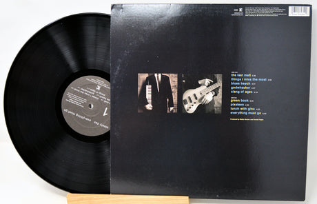 A vinyl record partially pulled from its black outer jacket, displayed on a small wooden stand against a white background. The back cover shows two small square photos and a track list printed in white and yellow text.