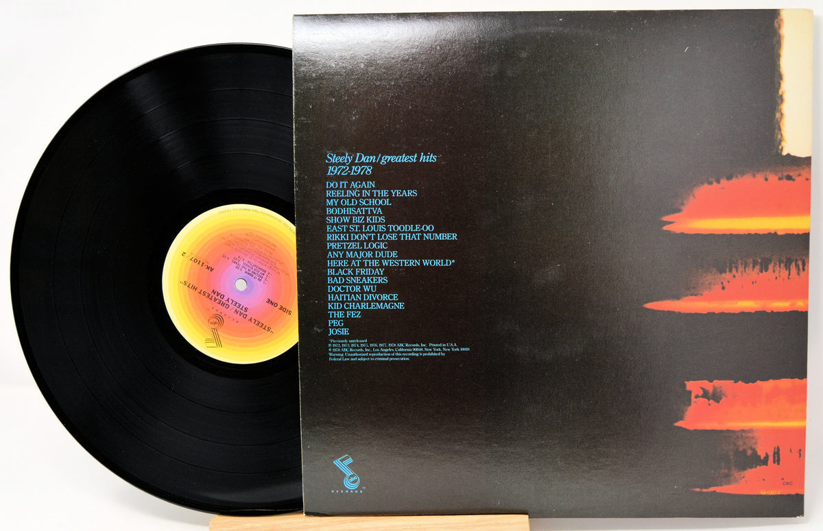 A vinyl record partially pulled from its sleeve, showing the black record with a yellow-orange center label and the back cover of the album in black with blue tracklisting text and orange brushstroke graphics.