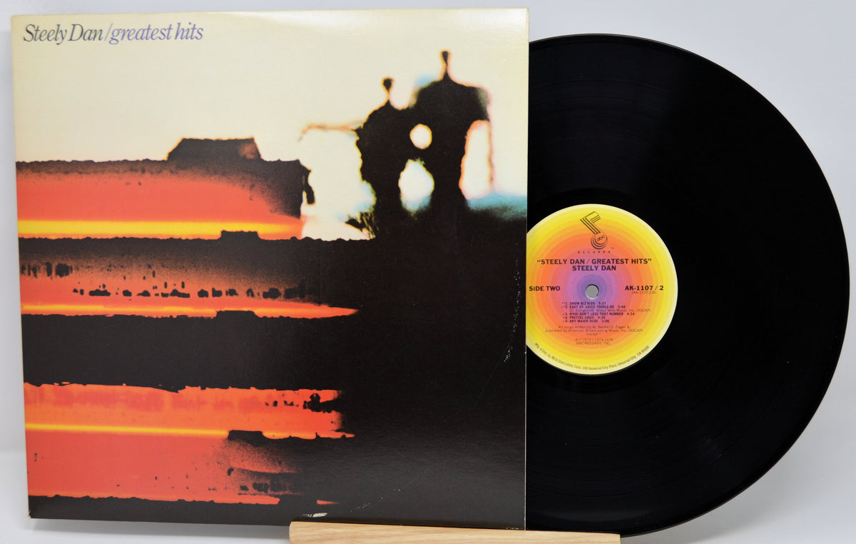 Steely Dan's 'Greatest Hits' vinyl record album with the LP partially pulled from the sleeve, showing the black record and a yellow-and-orange center label against a light gray background.