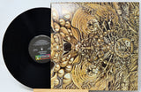 A vinyl record partially pulled from its illustrated album cover, showing the black record and the detailed gold-and-brown artwork on the sleeve against a light background.