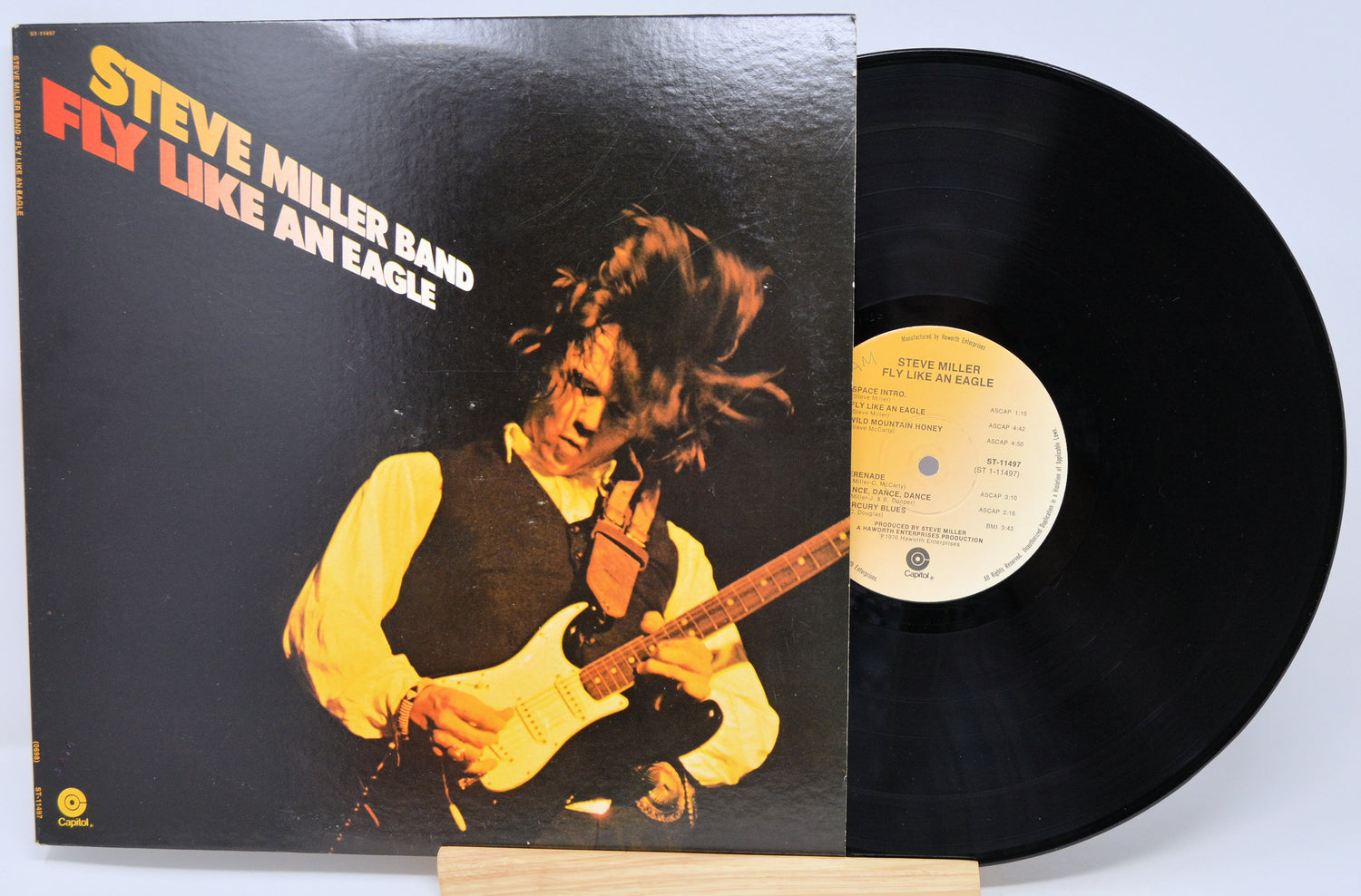 Steve Miller Band's 'Fly Like An Eagle' vinyl record LP, showing the black album cover with its artwork and the black vinyl record partially pulled out, displayed on a small wooden stand against a light gray background.