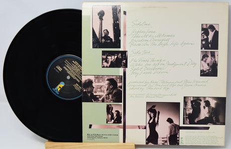 A vinyl record partially pulled from its printed album cover, displayed on a small wooden stand against a light gray background. The black record has a blue and yellow label, and the cover features a cream background with sepia-toned photographs and handwritten text.
