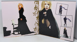 An open printed photo album or book spread, showing two pages with fashion photographs of a blonde woman in a long black dress. The left page has a pale pink background with ornate gold leaf decoration, and the right page contains multiple inset photos of the model wearing the dress from different angles.