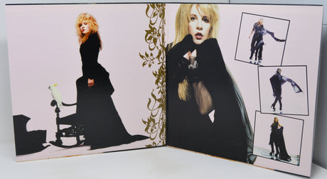 An open printed photo album or book spread, showing two pages with fashion photographs of a blonde woman in a long black dress. The left page has a pale pink background with ornate gold leaf decoration, and the right page contains multiple inset photos of the model wearing the dress from different angles.