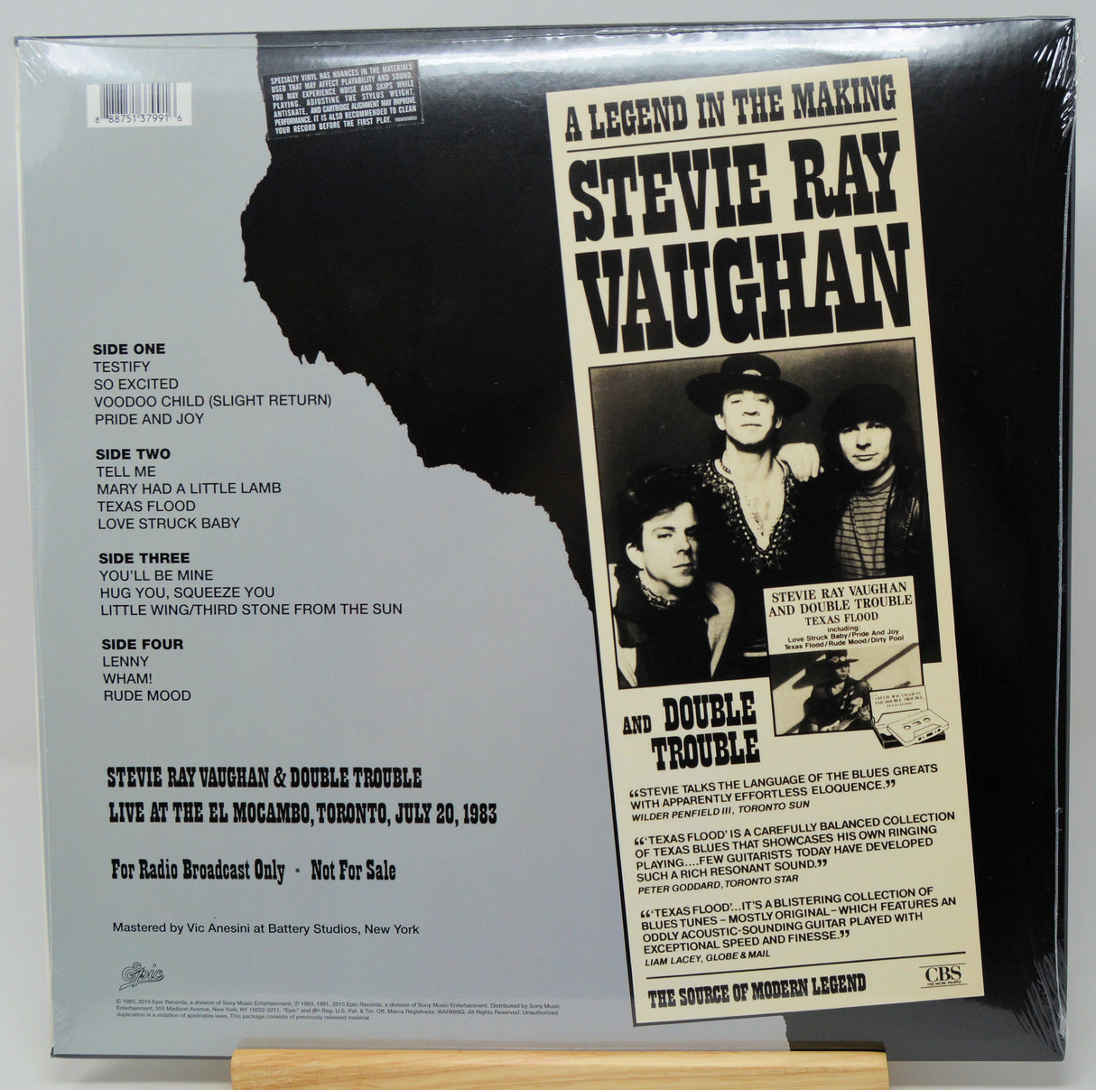 The back cover of a Stevie Ray Vaughan double-sided vinyl record, sealed in plastic shrink wrap, displayed on a wooden stand against a white background.