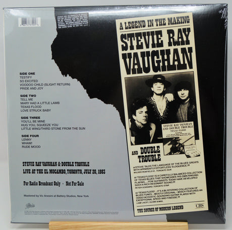 The back cover of a Stevie Ray Vaughan double-sided vinyl record, sealed in plastic shrink wrap, displayed on a wooden stand against a white background.