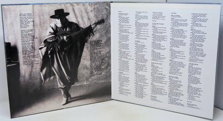 An open vinyl record sleeve or booklet showing a black-and-white cover photo of a person in a hat and coat holding a guitar, with dense printed text on the right page.