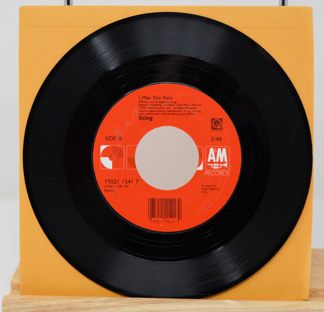 A vinyl record of Sting's song 'All This Time' with a visible orange and white label, placed on a yellow surface.