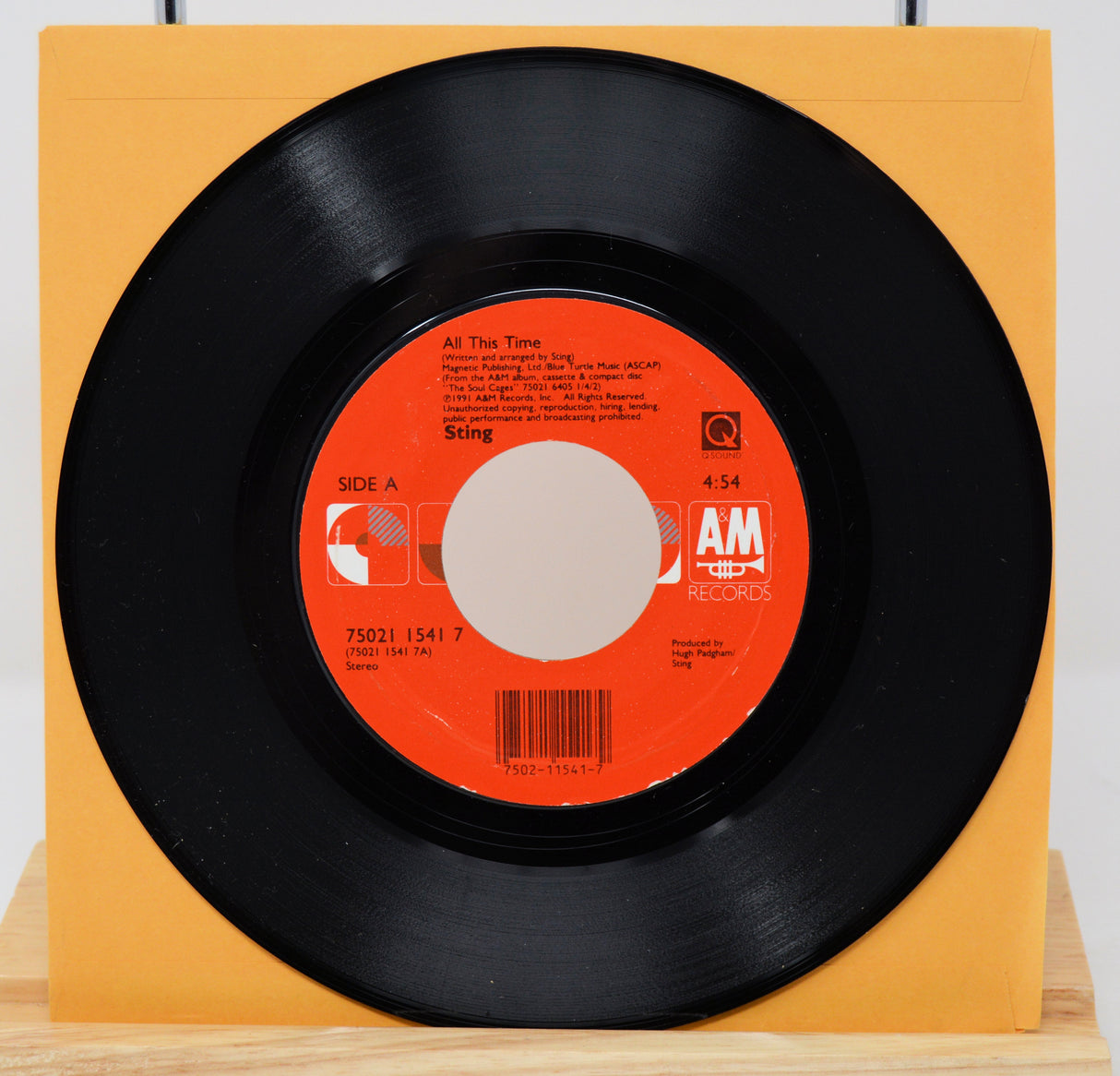 A vinyl record of Sting's song 'All This Time' with a visible orange and white label, placed on a yellow surface.