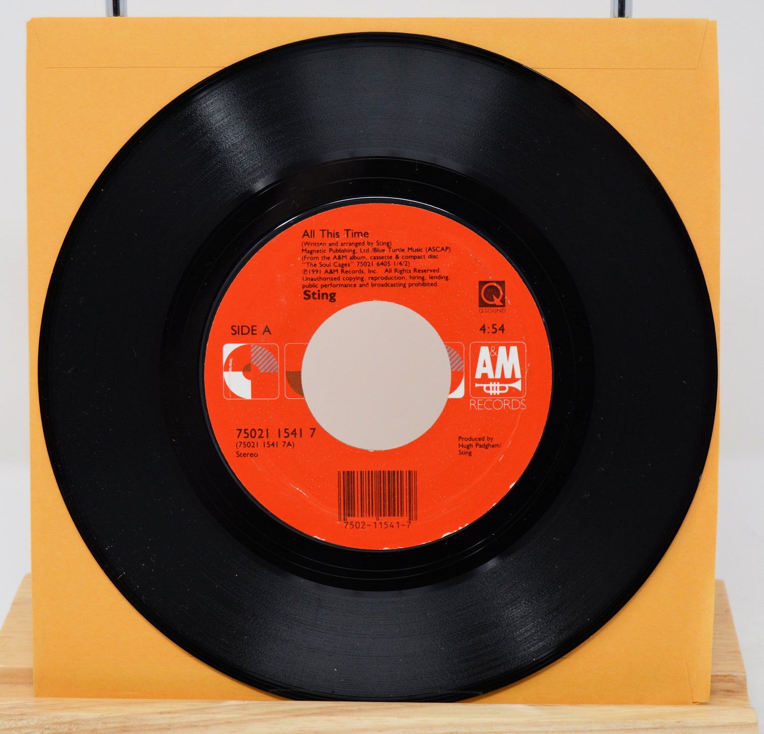 A vinyl record of Sting's song 'All This Time' with a visible orange and white label, placed on a yellow surface.