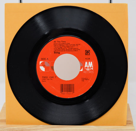 A vinyl record of Sting's song 'All This Time' with a visible orange and white label, placed on a yellow surface.