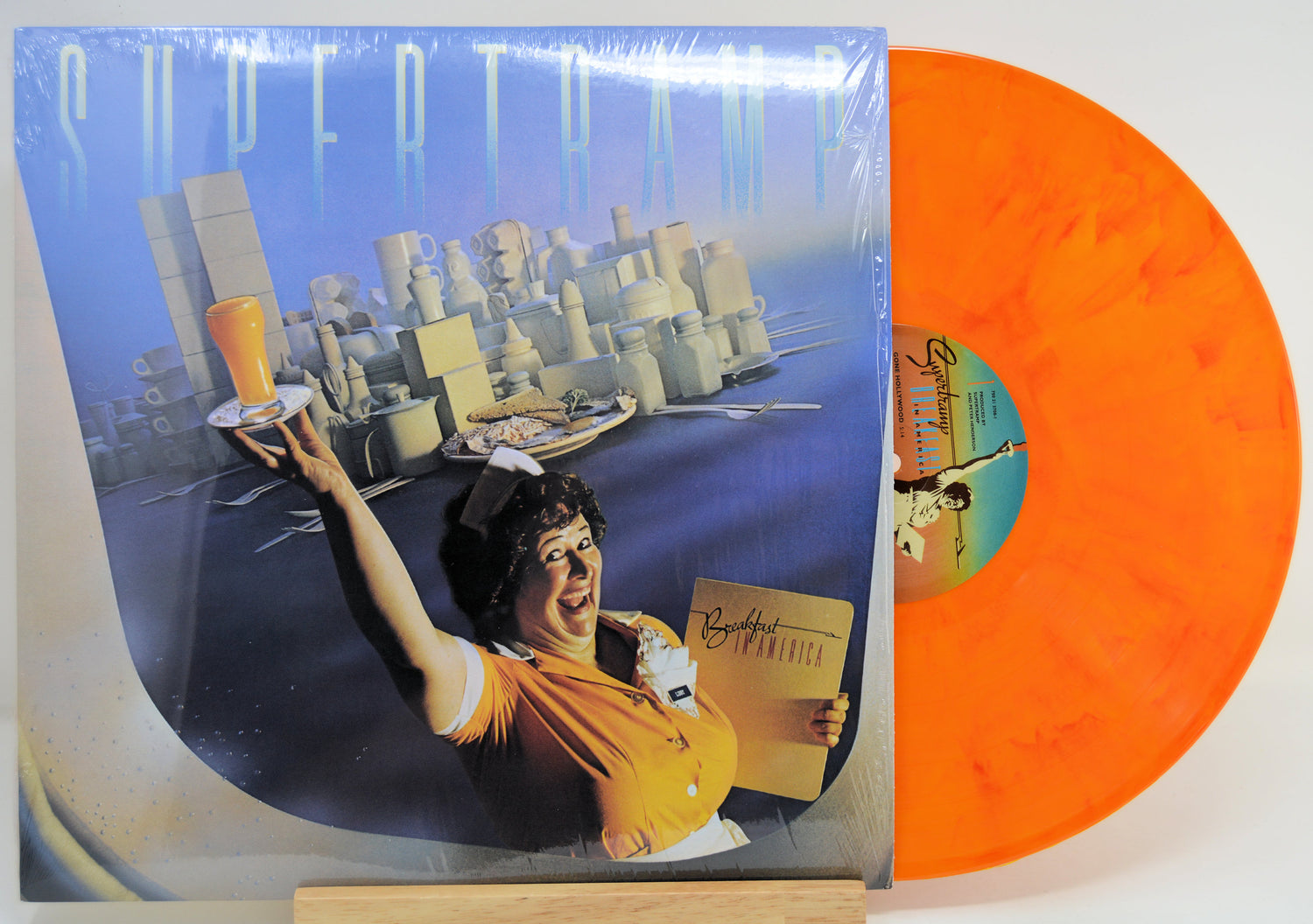 A vinyl record album with its cover jacket visible on the left and the orange marbled record partially pulled out on the right. The cover artwork shows a surreal cityscape and a woman in a yellow top holding a drink and a menu.