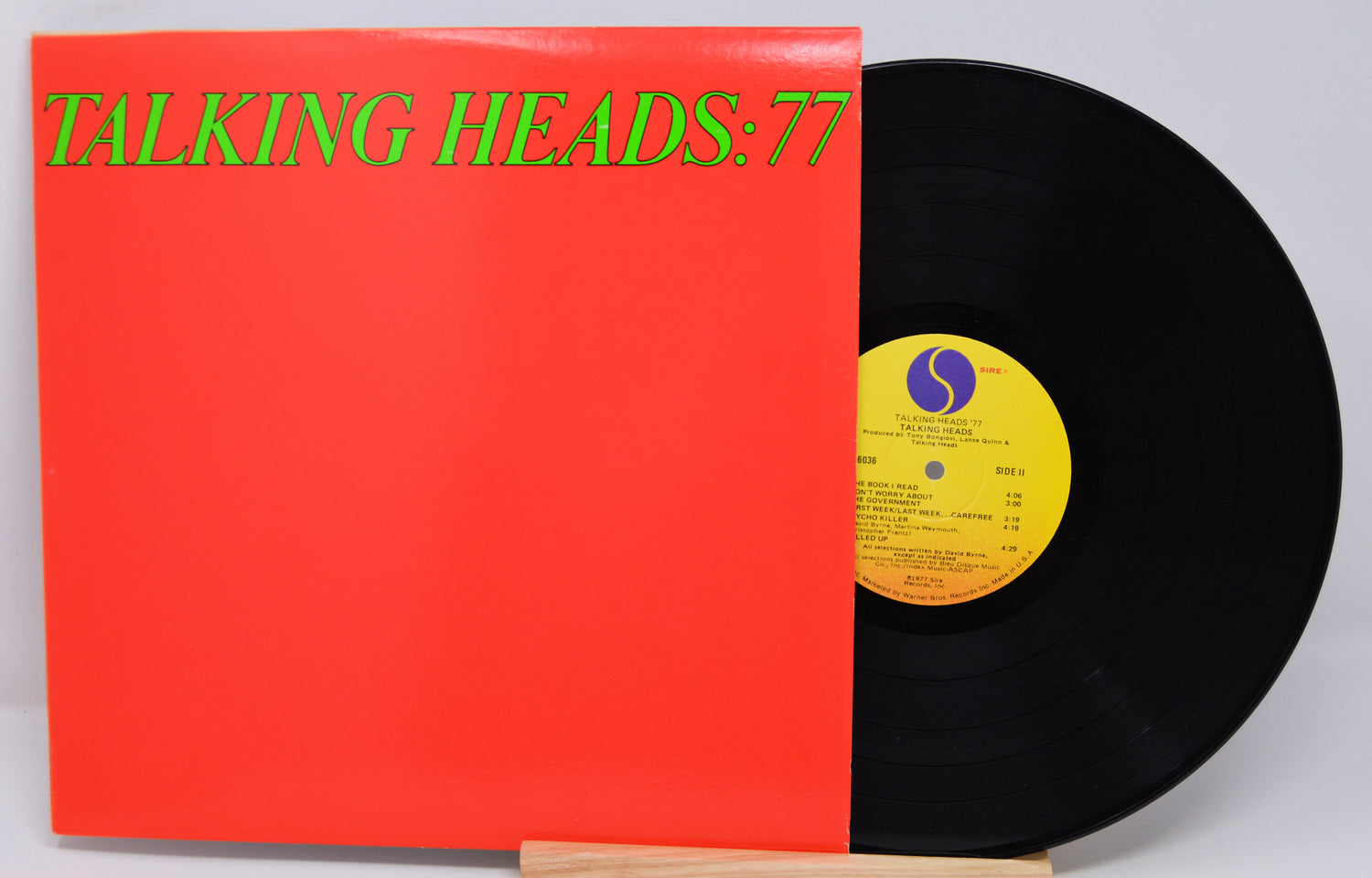 A vinyl record titled 'Talking Heads - 77' with a red cover and a black record inside. The label shows the band name and the album title.