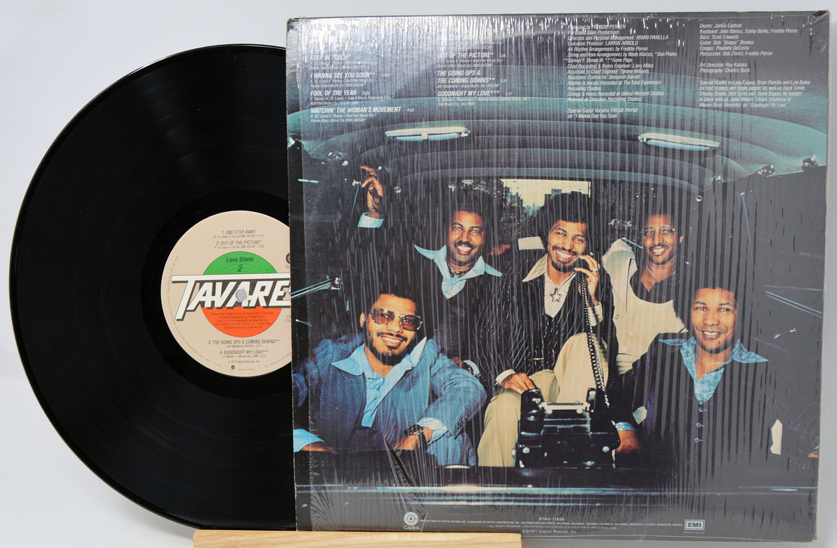 A vinyl record partially pulled from its plastic-wrapped album cover, showing the black record on the left and the cover art on the right. The center label reads Tavares.