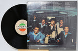 A vinyl record partially pulled from its plastic-wrapped album cover, showing the black record on the left and the cover art on the right. The center label reads Tavares.