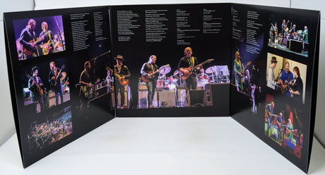 A large printed triptych display or poster featuring multiple concert photographs of a band performing, with text blocks printed on the black background panels, photographed on a white surface against a white wall.