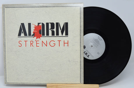 A vinyl record titled 'The Alarm - Strength' with a record cover featuring the band's name and the album title in red and black text on a white background, and a black vinyl record inside.