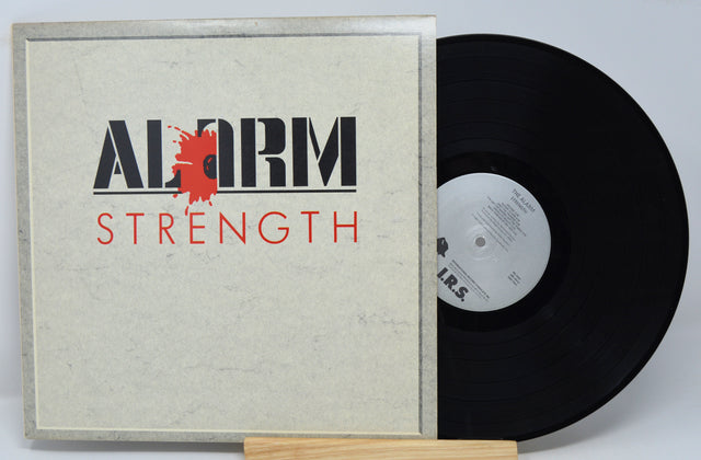 A vinyl record titled 'The Alarm - Strength' with a record cover featuring the band's name and the album title in red and black text on a white background, and a black vinyl record inside.