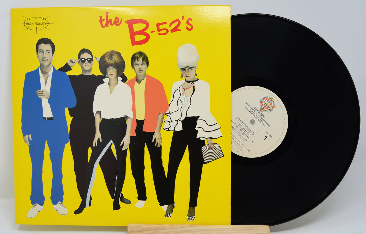 The image shows a vinyl record of 'The B-52's' with a yellow album cover featuring black and white band photo. The record is placed on a wooden platter.