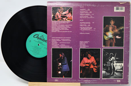 A vinyl record partially pulled from its back cover sleeve, showing the black record, a green center label, and the purple back cover with printed track listings and performer photos.