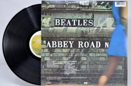 Beatles 'Abbey Road' vinyl record partially pulled from its cover, showing the black LP disc and the front cover artwork with a blurred figure in blue on a brick wall background.