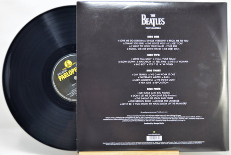 The Beatles - Past Masters vinyl record shown with the black LP partially pulled from the back of the outer paper sleeve. The sleeve displays the band's name and track listing in white text on a black background.